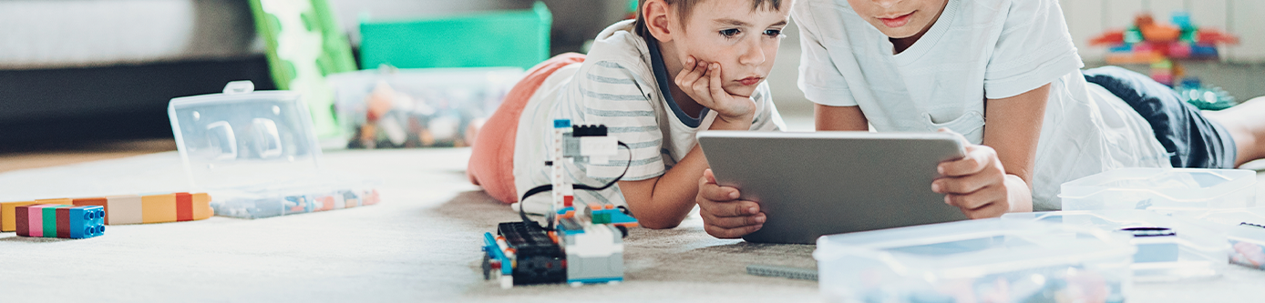 Two young brothers laying on the living room floor surrounded by toys and playing on their tablet.