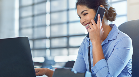 A business woman speaking on a office phone and working on a laptop at her desk.