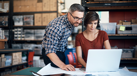 A man and women reviewing information on laptop at a desk within a manufacturing facility.