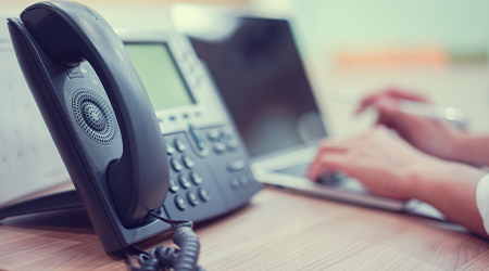 A close up image of a business phone and man working on a laptop.