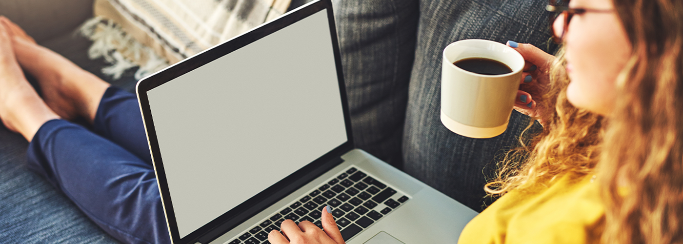 A women laying on her couch enjoying a cup of coffee and using her laptop.